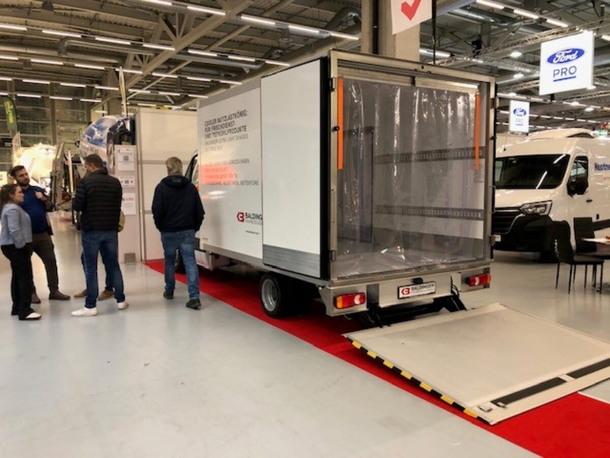 Messe mit Besuchern und seitlicher Heckansicht des Baldinger Fahrzeugbau Kühltransport Vorführwagen offen mit abgesenkter Hebebühne und Isoliervorhang im Hintergrund der neue Renault Master Kühltransporter
